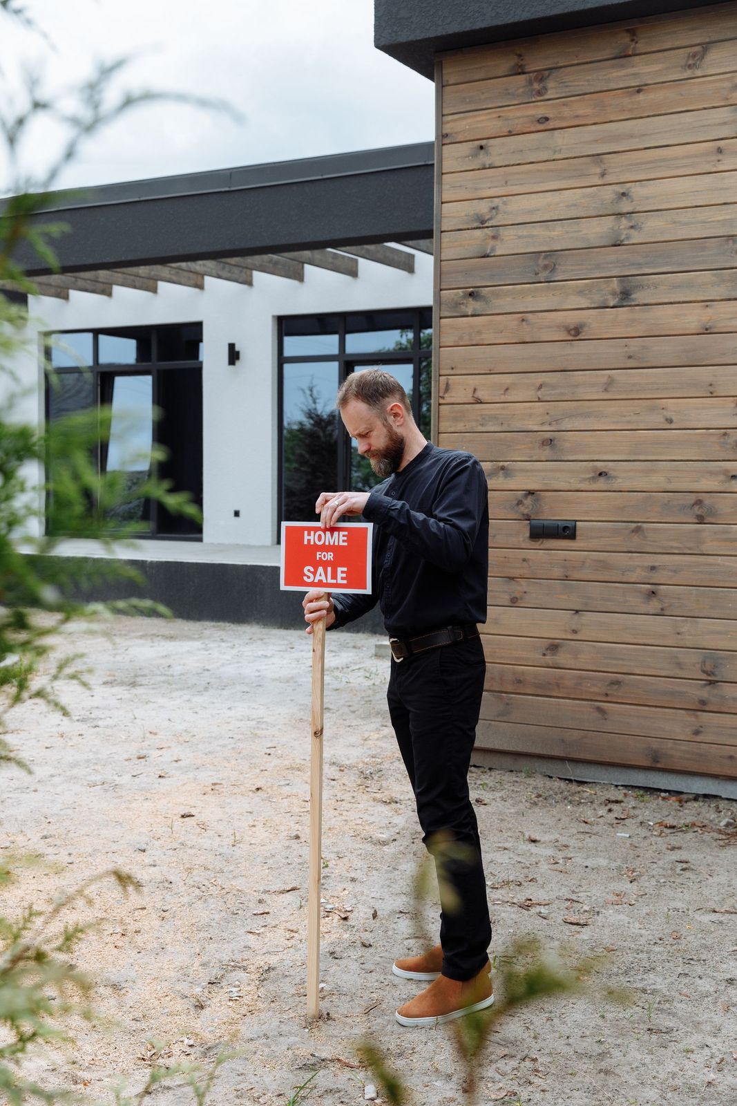 A man fixing a Home for Sale board into the gravel outside a timber-clad house